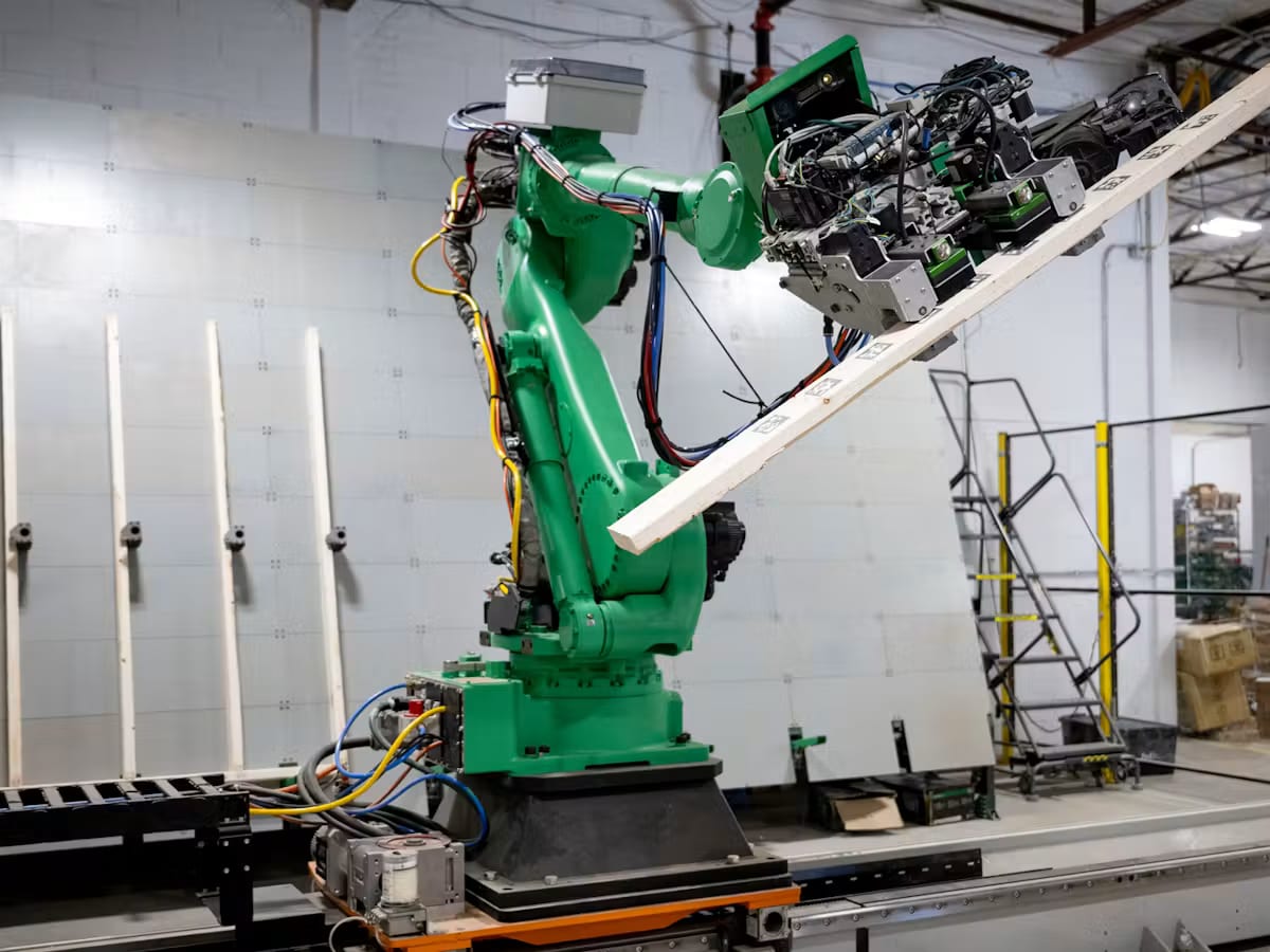A green robotic arm lifting a lumber stud onto a wall panel on the Reframe Systems microfactory line